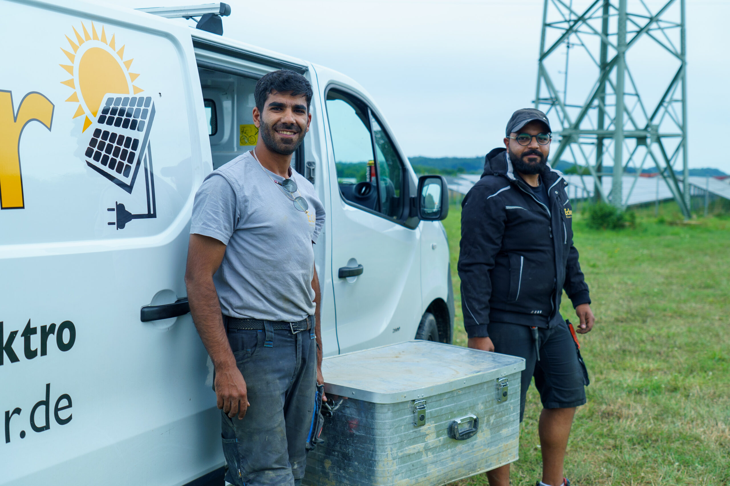 Zwei Mitarbeiter von Elektro Ecker halten gemeinsam eine Kiste. Sie stehen auf einer Wiese. Im Hintergrund ist ein Arbeitsfahrzeug der Firma.